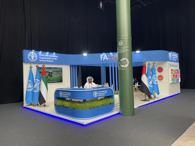 Wider view of the FAO exhibition stand — reception desk with grass element, UAE and FAO flag arrangement, agriculture-themed video screen, and SDG goals graphic